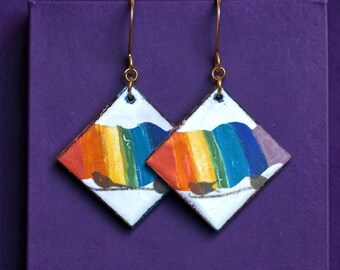 May include: A pair of diamond-shaped dangle earrings featuring a white background with a vibrant rainbow-colored arch design. Each earring displays a small black bird silhouette perched on a thin line at the bottom, suspended by gold-toned hooks.