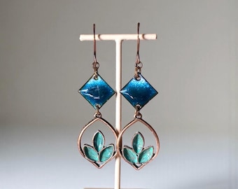May include: A pair of copper-colored dangle earrings. Each earring features a teal, leaf-like design within a teardrop shape, topped with a blue diamond. The earrings are displayed on a small, light-colored stand.