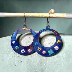 Puede incluir: Un par de pendientes de aro circulares con un acabado metálico azul profundo iridiscente y textura moteada. Cada aro está decorado con pequeños cabochones con motivos florales en tonos blanco, rosa, amarillo y azul claro.