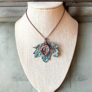 May include: A copper necklace with a turquoise patina featuring a large leaf pendant with a smaller heart-shaped pendant. The necklace is on a white fabric display stand.