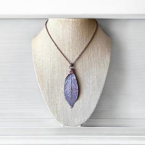 May include: A copper necklace with a leaf-shaped pendant. The pendant is a light purple colour with a textured surface. The necklace is on a mannequin.