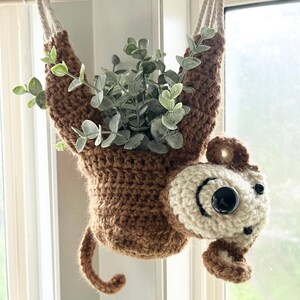 May include: A crocheted monkey planter hanging from a wooden dowel. The monkey is brown with a white face and black eyes. The planter is filled with a green plant.