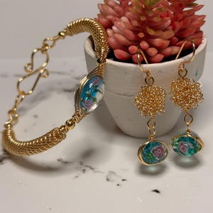 Puede incluir: Una pulsera de alambre dorado y un par de pendientes a juego. Cada pieza presenta una cuenta de vidrio transparente con un diseño floral azul y rosa. La pulsera tiene un cierre de gancho y los pendientes tienen racimos de alambre dorado.
