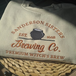 May include: A light green sweatshirt with an embroidered logo that says "Sanderson Sisters Brewing Co. Premium Witch's Brew" with an image of a cauldron and the text "Est. 1698".