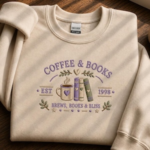 Puede incluir: Una sudadera beige de cuello redondo con un diseño bordado que incluye el texto COFFEE & BOOKS, EST 1998 y BREWS, BOOKS & BLISS. El gráfico muestra una taza de café, tres libros apilados, corazones pequeños y ramas con hojas en morado y verde.
