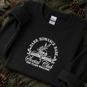 May include: A black crewneck sweatshirt featuring a white graphic print of an open book, a skull, a rose, and a dagger. The text reads: DARK ROMANCE BABE SOCIAL CLUB RED FLAGS WELCOME. The garment has a Gildan brand label at the inner neckline.