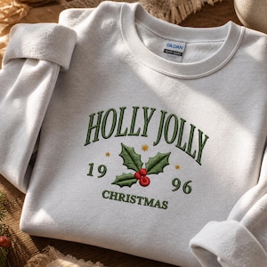 Può includere: Una felpa girocollo grigio chiaro con il testo ricamato HOLLY JOLLY CHRISTMAS in verde scuro. Il design include un rametto di agrifoglio centrale con foglie verdi e bacche rosse, affiancato dai numeri 19 e 96 in colore verde.