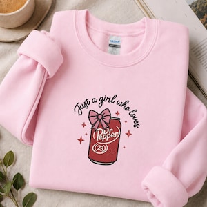 May include: A light pink crewneck sweatshirt featuring an embroidered design of a red soda can topped with a pink bow. The text "Just a girl who loves" is arched above the can, surrounded by small embroidered star accents in a dark thread.