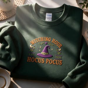 May include: A dark green crewneck sweatshirt featuring an embroidered design with a purple witch hat, a yellow buckle, and the text "WITCHING HOUR" and "HOCUS POCUS EST 1998" in orange. The design includes small stars, crescent moons, and floral accents.