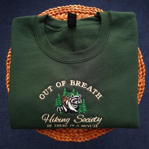 K&ouml;nnte beinhalten: Dunkelgr&uuml;nes Sweatshirt mit gesticktem Text und einer Waschb&auml;r-Grafik. Der Text lautet "OUT OF BREATH Hiking Society BE THERE IN A MINUTE". Der Waschb&auml;r tr&auml;gt einen Rucksack und ist von gr&uuml;nen B&auml;umen umgeben.