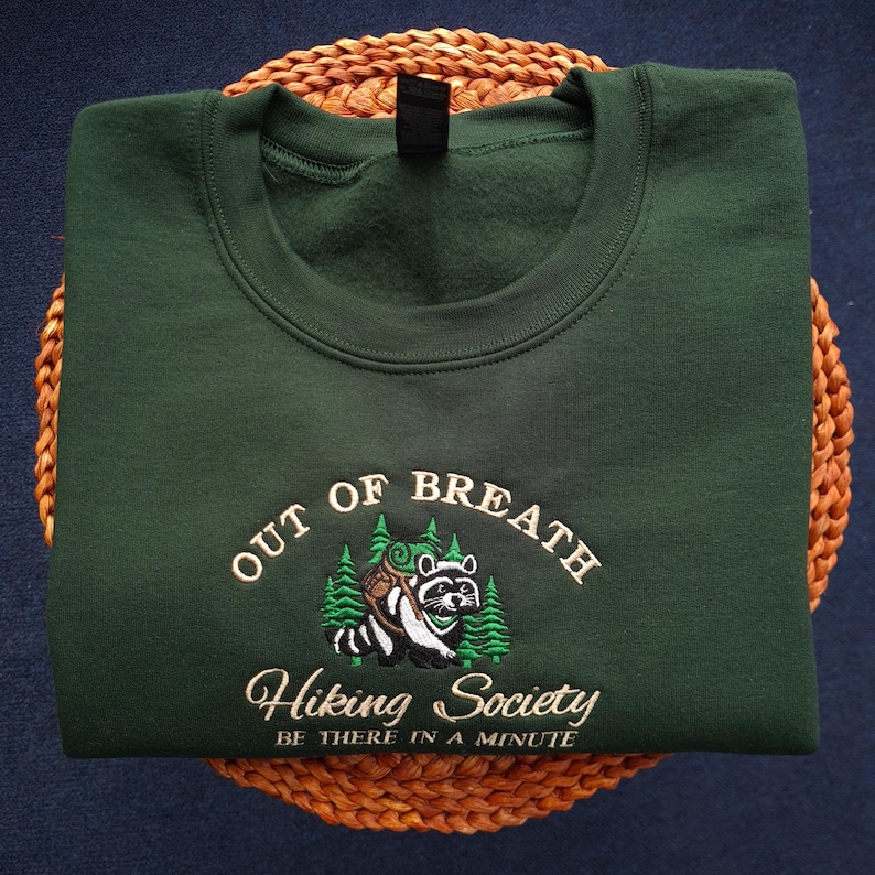K&ouml;nnte beinhalten: Dunkelgr&uuml;nes Sweatshirt mit gesticktem Text und einer Waschb&auml;r-Grafik. Der Text lautet "OUT OF BREATH Hiking Society BE THERE IN A MINUTE". Der Waschb&auml;r tr&auml;gt einen Rucksack und ist von gr&uuml;nen B&auml;umen umgeben.