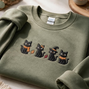 Puede incluir: Una sudadera de cuello redondo color verde oliva con un dise&ntilde;o bordado de cuatro gatos negros. Los gatos lucen accesorios naranjas que incluyen una bufanda, un lazo, un sombrero de bruja y un cuello con volantes, junto a peque&ntilde;as hojas naranjas y rojas.