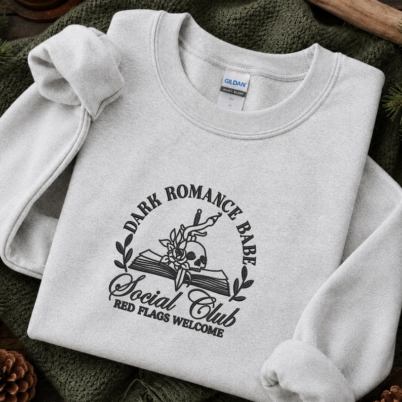 May include: A light grey crewneck sweatshirt featuring a black embroidered graphic on the chest. The design includes an open book, a skull, a rose, and a dagger, surrounded by the text Dark Romance Babe Social Club Red Flags Welcome.