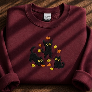 May include: A maroon long-sleeve crewneck sweatshirt featuring an embroidered design of three black cats with large yellow eyes playing among scattered orange and yellow autumn leaves. The inner neck label displays the brand name Gildan.