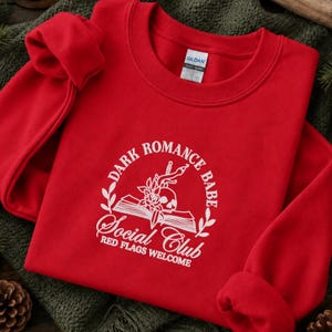 May include: A bright red crewneck sweatshirt featuring a white graphic print. The design includes an open book, a skull, a rose, and a dagger, surrounded by the text "DARK ROMANCE BABE SOCIAL CLUB" and "RED FLAGS WELCOME" in a circular layout.