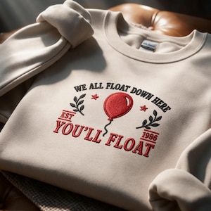 May include: A beige crewneck sweatshirt featuring embroidered text that reads WE ALL FLOAT DOWN HERE and YOU'LL FLOAT. The design includes a red balloon, two black leafy branches, two small red stars, and the text EST 1986 in red embroidery.