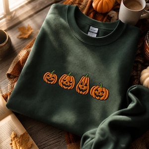 Pode incluir: Uma camisola de gola redonda em verde floresta escuro com um design bordado de quatro abóboras cor de laranja com expressões faciais esculpidas. A peça inclui uma etiqueta da marca Gildan na parte interior da gola.