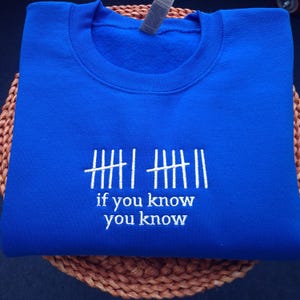 May include: A royal blue sweatshirt with white embroidered text and tally marks. The text reads "if you know you know". The sweatshirt is folded and resting on a woven surface.