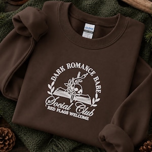 May include: A dark brown crewneck sweatshirt featuring a white graphic print of an open book, a skull, a rose, and a dagger. The text reads: DARK ROMANCE BABE SOCIAL CLUB RED FLAGS WELCOME. The garment has a Gildan brand label at the inner neckline.