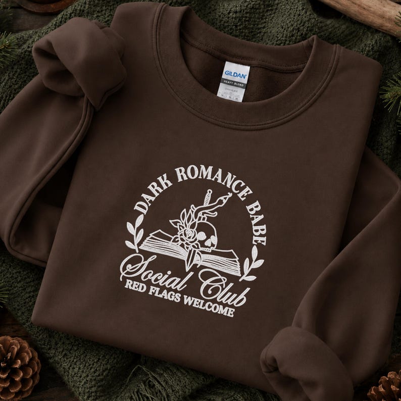 May include: A dark brown crewneck sweatshirt featuring a white graphic print of an open book, a skull, a rose, and a dagger. The text reads: DARK ROMANCE BABE SOCIAL CLUB RED FLAGS WELCOME. The garment has a Gildan brand label at the inner neckline.
