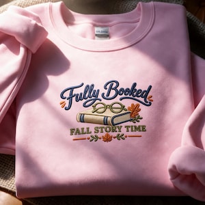 Puede incluir: Una sudadera de cuello redondo rosa claro con un diseño bordado que incluye el texto "Fully Booked" en azul oscuro y "FALL STORY TIME" en verde oliva. El gráfico muestra un libro beige, gafas redondas y hojas de otoño naranjas y verdes.