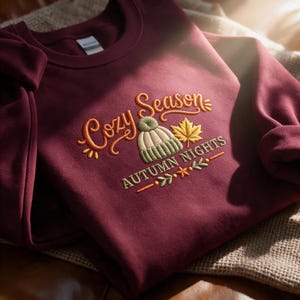 May include: A maroon crewneck sweatshirt featuring embroidered text that reads Cozy Season and AUTUMN NIGHTS. The design includes a light green knit beanie, a yellow maple leaf, and small decorative leaf accents in orange and green colours.