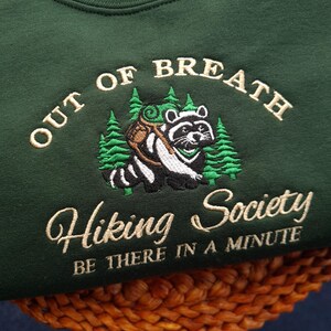 K&ouml;nnte beinhalten: Dunkelgr&uuml;nes Sweatshirt mit gesticktem Text und Waschb&auml;r-Design. Der Text lautet "OUT OF BREATH Hiking Society BE THERE IN A MINUTE". Der Waschb&auml;r tr&auml;gt einen Rucksack und ist von gr&uuml;nen B&auml;umen umgeben.