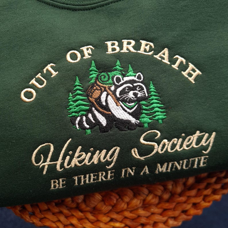 K&ouml;nnte beinhalten: Dunkelgr&uuml;nes Sweatshirt mit gesticktem Text und Waschb&auml;r-Design. Der Text lautet "OUT OF BREATH Hiking Society BE THERE IN A MINUTE". Der Waschb&auml;r tr&auml;gt einen Rucksack und ist von gr&uuml;nen B&auml;umen umgeben.