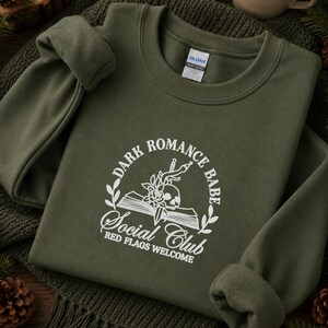 May include: An olive green crewneck sweatshirt featuring a white embroidered graphic of an open book, a skull, a rose, and a dagger. The text reads: Dark Romance Babe Social Club, Red Flags Welcome. The garment has a Gildan brand neck label.