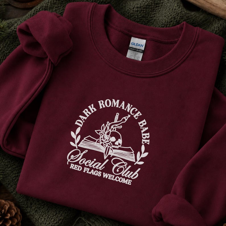 May include: A maroon crewneck sweatshirt featuring white embroidery. The design includes an open book, a skull, a rose, and a dagger, surrounded by the text "DARK ROMANCE BABE SOCIAL CLUB" and "RED FLAGS WELCOME" in a circular arrangement.