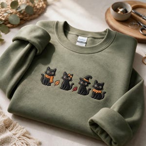 Puede incluir: Una sudadera de cuello redondo color verde oliva con un dise&ntilde;o bordado de cuatro gatos negros. Los gatos aparecen con varios accesorios, incluyendo una bufanda naranja, un lazo peque&ntilde;o, un sombrero de bruja y un cuello con volantes, junto a dos hojas.