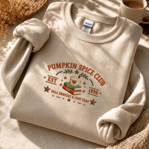 Peut inclure: Un sweat-shirt beige à col rond avec une broderie indiquant PUMPKIN SPICE CLUB, EST 1998, et FALL BREEZES & COZY TEAS. Le motif comprend une pile de deux livres, une tasse avec un cœur, des étoiles et du feuillage en orange et vert.