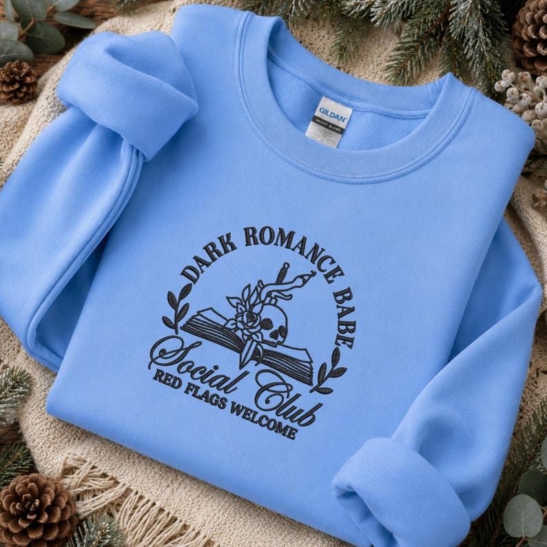 May include: A light blue crewneck sweatshirt featuring black embroidery on the chest. The design includes an open book, a skull, a dagger, and floral elements, with the text "DARK ROMANCE BABE SOCIAL CLUB RED FLAGS WELCOME" in a circular layout.