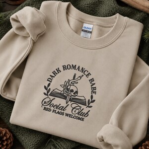 May include: A beige crewneck sweatshirt featuring black embroidery on the chest. The design includes an open book, a skull, a rose, and a dagger, surrounded by the text "DARK ROMANCE BABE SOCIAL CLUB" and "RED FLAGS WELCOME" in a circular layout.