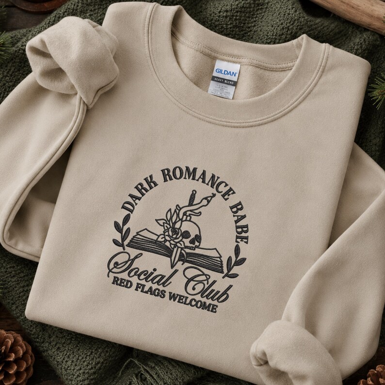 May include: A beige crewneck sweatshirt featuring black embroidery on the chest. The design includes an open book, a skull, a rose, and a dagger, surrounded by the text "DARK ROMANCE BABE SOCIAL CLUB" and "RED FLAGS WELCOME" in a circular layout.