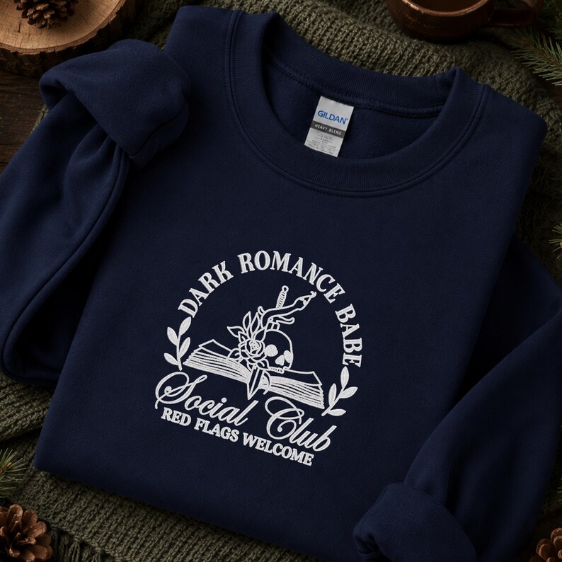 May include: A navy blue crewneck sweatshirt featuring a white graphic print. The design includes an open book, a skull, a rose, and a dagger, surrounded by the text "DARK ROMANCE BABE SOCIAL CLUB" and "RED FLAGS WELCOME" in a circular layout.