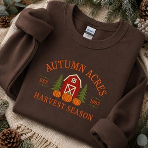 May include: A dark brown crewneck sweatshirt featuring an embroidered graphic of a red barn, two green pine trees, and three orange pumpkins. The text reads "AUTUMN ACRES" in an arch, "EST. 1987" on the sides, and "HARVEST SEASON" below.