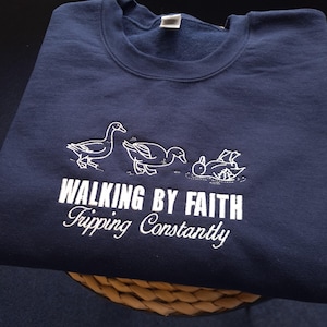 Puede incluir: Sudadera azul marino con patos bordados en blanco y el texto "WALKING BY FAITH Tripping Constantly". La sudadera está doblada sobre una cesta tejida. El diseño presenta tres patos en varias poses.