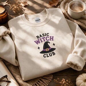 May include: A cream-colored crewneck sweatshirt featuring an embroidered design on the chest. The embroidery includes the text "BASIC WITCH CLUB" in black and purple, centered above and below a black witch hat with a purple band and gold buckle.
