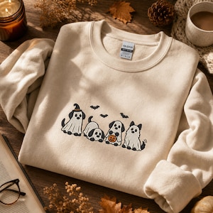 May include: A beige crewneck sweatshirt featuring an embroidered design of four dogs dressed as ghosts for Halloween. One dog wears a witch hat, and another holds a small orange jack-o'-lantern bucket. Three small black bats are embroidered above.