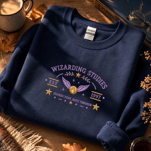 Könnte beinhalten: Ein marineblaues Sweatshirt mit Rundhalsausschnitt und einer Stickerei mit der Aufschrift WIZARDING STUDIES, EST 1997 sowie LEARNING TODAY, MAGIC TOMORROW. Das Design zeigt einen goldenen geflügelten Ball, gelbe Sterne und violette Akzente.