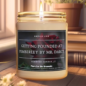 May include: A scented candle with a gold lid and a black label that reads "Smells Like Getting Pounded at Pemberley by Mr. Darcy". The label also says "Scented Candle Flare for the Dramatic Vegan Soy Candle | Cotton Wick | Ethically Sourced and Made"