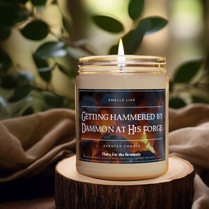 以下が含まれることがあります： 白いラベルに「Smells Like Getting Hammered by Damon at His Forge」と書かれた茶色の大豆ろうそく。ラベルには「Scented Candle Flare for the Dramatic Vegan Soy Candle | Cotton Wick | Ethically Sourced and Made」というテキストも含まれています