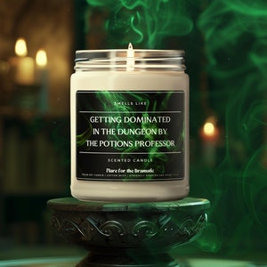May include: A white soy candle with a black label that reads "Smells Like Getting Dominated in the Dungeon by the Potions Professor" and "Scented Candle Flare for the Dramatic". The candle is on a dark wooden pedestal with a green smoke-like background.