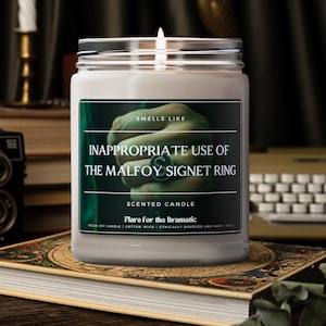 May include: A white candle jar with a green label that reads "Smells Like Inappropriate Use of the Malfoy Signet Ring" and "Scented Candle Flare for the Dramatic". The label also includes the text "Vegan Soy Candle | Cotton Wick | Ethically Sourced and Made | 11 oz."