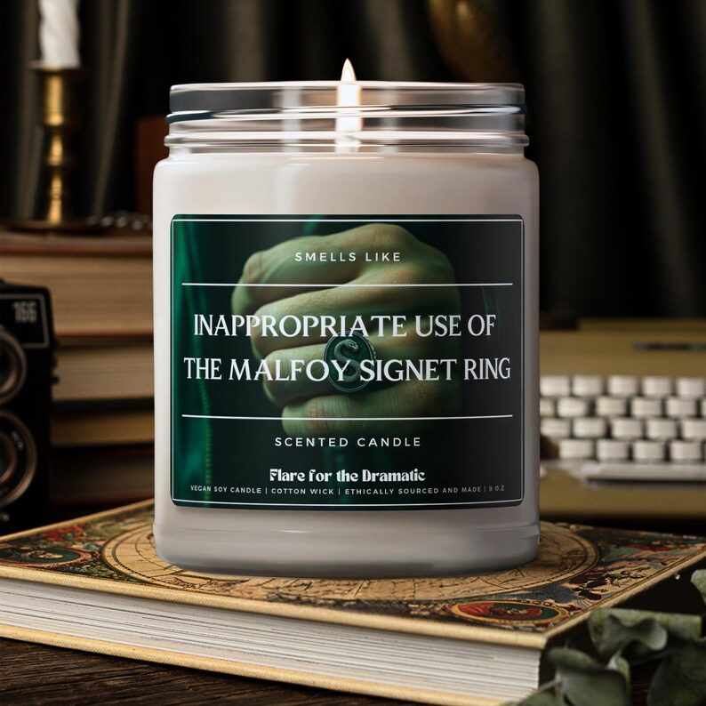 May include: A white candle jar with a green label that reads "Smells Like Inappropriate Use of the Malfoy Signet Ring" and "Scented Candle Flare for the Dramatic". The label also includes the text "Vegan Soy Candle | Cotton Wick | Ethically Sourced and Made | 11 oz."