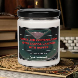May include: A white candle jar with a black lid. The label reads "Smells Like Coffee and Contemplation After Carnal Caresses With Hopper" and "Scented Candle Flare for the Dramatic".