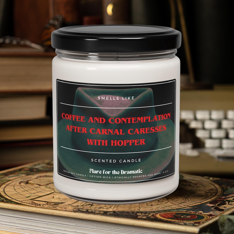 May include: A white candle jar with a black lid. The label reads "Smells Like Coffee and Contemplation After Carnal Caresses With Hopper" and "Scented Candle Flare for the Dramatic".