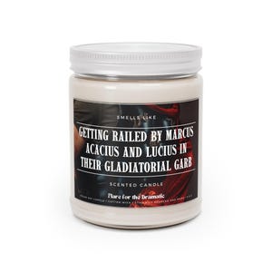 May include: A white candle jar with a silver lid. The label reads "Smells Like Getting Railed by Marcus Acacius and Lucius in Their Gladiatorial Garb". The candle is labelled "Scented Candle Flare for the Dramatic" and is a pecan soy candle with a cotton wick.