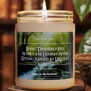 Könnte beinhalten: Eine durchsichtige Glas-Kerze in einem Glas mit einem beigefarbenen Etikett, auf dem "Smells Like Being Tenderly Fed After-Care Lembas After Getting Railed By Legolas" und "Scented Candle Flare for the Dramatic" steht. Die Kerze ist angezündet und die Flamme ist sichtbar.